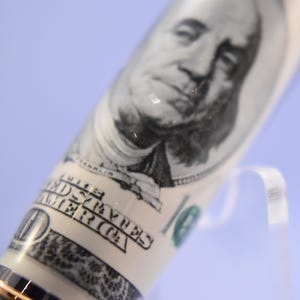 May include: A close-up of a pen with a design resembling a United States $100 bill. The pen's barrel shows Benjamin Franklin's portrait and the words "The United States of America."