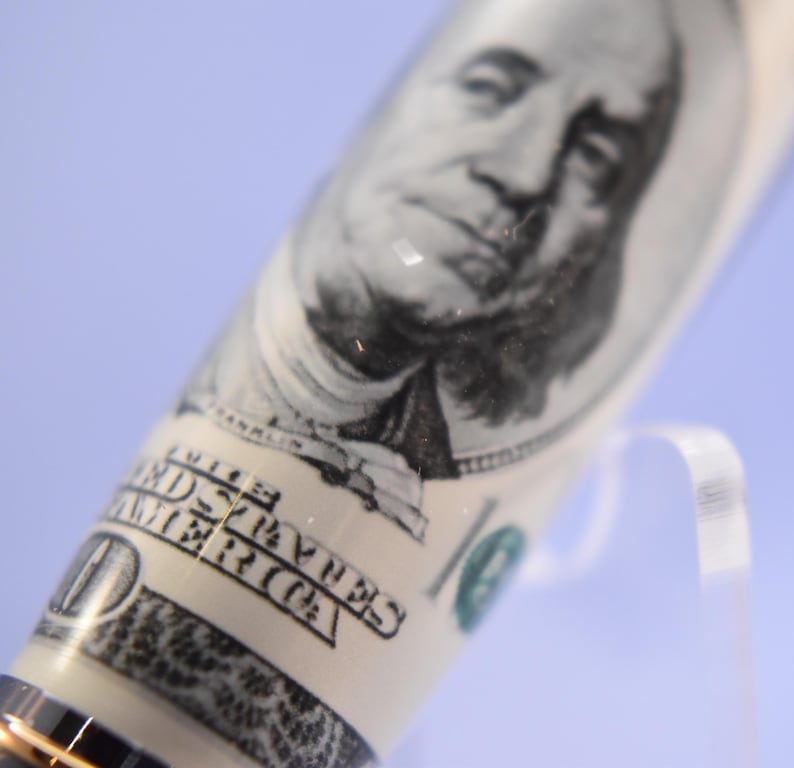 May include: A close-up of a pen with a design resembling a United States $100 bill. The pen's barrel shows Benjamin Franklin's portrait and the words "The United States of America."