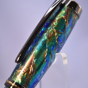 K&ouml;nnte beinhalten: Nahaufnahme eines Stifts mit schimmerndem, irisierendem Finish in Blau-, Gr&uuml;n- und Goldt&ouml;nen. Der Stift hat einen schwarzen Clip und silberne Akzente. Der Stift wird auf einem transparenten St&auml;nder pr&auml;sentiert.