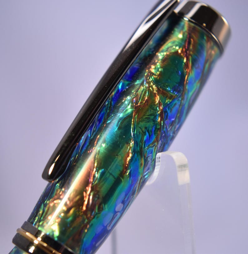 K&ouml;nnte beinhalten: Nahaufnahme eines Stifts mit schimmerndem, irisierendem Finish in Blau-, Gr&uuml;n- und Goldt&ouml;nen. Der Stift hat einen schwarzen Clip und silberne Akzente. Der Stift wird auf einem transparenten St&auml;nder pr&auml;sentiert.