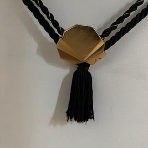 May include: A gold-colored, fan-shaped pendant with a black tassel hangs from two black twisted cords with gold accents. The pendant is the focal point, with the tassel hanging below.