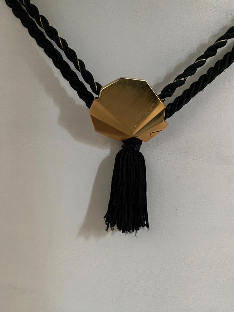May include: A gold-colored, fan-shaped pendant with a black tassel hangs from two black twisted cords with gold accents. The pendant is the focal point, with the tassel hanging below.