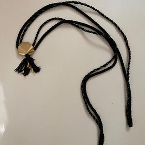 May include: A black cord necklace with a gold-colored geometric pendant and a small tassel. The cord is twisted with a silver thread. The necklace is displayed on a white surface.