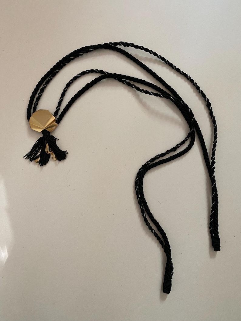 May include: A black cord necklace with a gold-colored geometric pendant and a small tassel. The cord is twisted with a silver thread. The necklace is displayed on a white surface.
