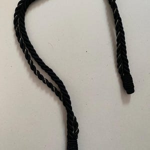 May include: Close-up of a black rope with a silver metallic thread woven throughout. The rope is twisted and has a decorative end. The background is a plain white surface. The rope is likely used for decorative purposes.