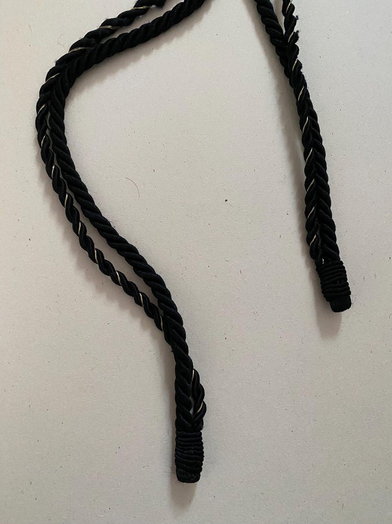 May include: Close-up of a black rope with a silver metallic thread woven throughout. The rope is twisted and has a decorative end. The background is a plain white surface. The rope is likely used for decorative purposes.