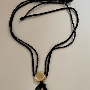 May include: A black cord necklace with a gold-colored geometric pendant and a black tassel. The necklace has a double-strand design and a simple, elegant aesthetic. The pendant is a multi-faceted shape.
