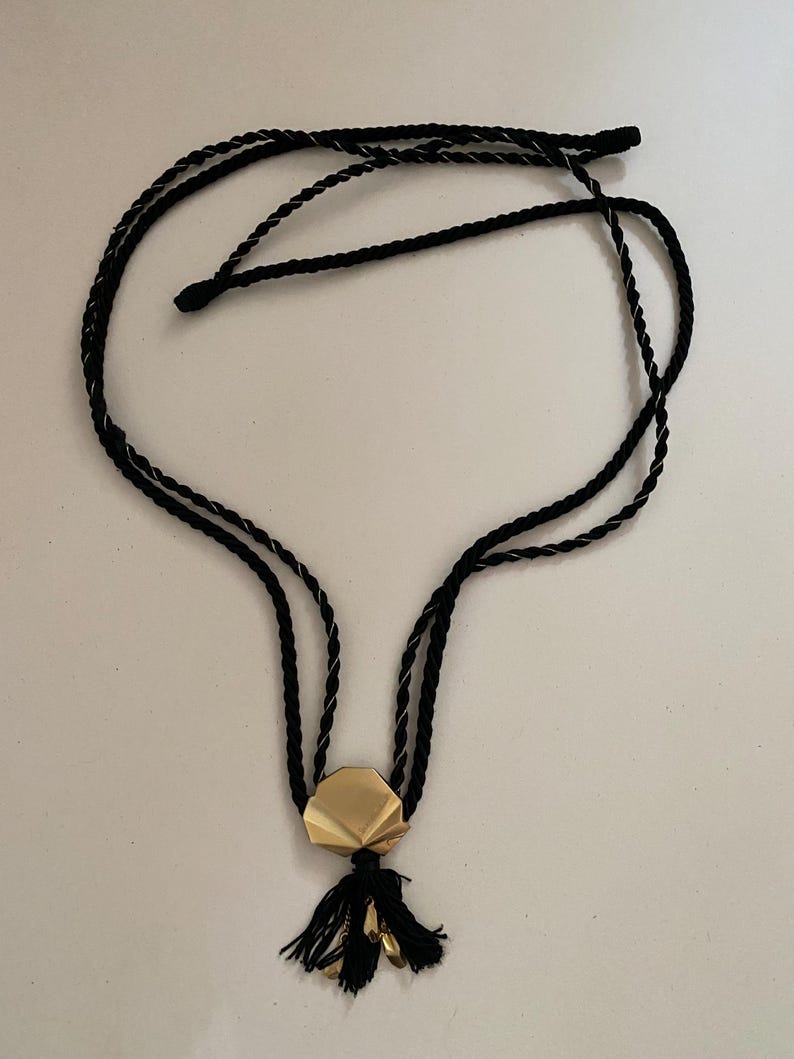 May include: A black cord necklace with a gold-colored geometric pendant and a black tassel. The necklace has a double-strand design and a simple, elegant aesthetic. The pendant is a multi-faceted shape.