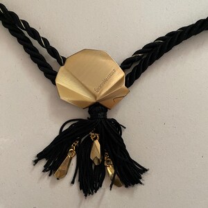 May include: A black necklace with a gold-colored pendant and tassel. The pendant is a geometric shape with the text "Gianni Versace" engraved on it. The tassel has gold-colored charms.