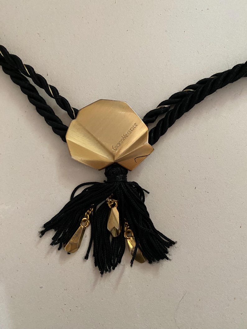 May include: A black necklace with a gold-colored pendant and tassel. The pendant is a geometric shape with the text "Gianni Versace" engraved on it. The tassel has gold-colored charms.