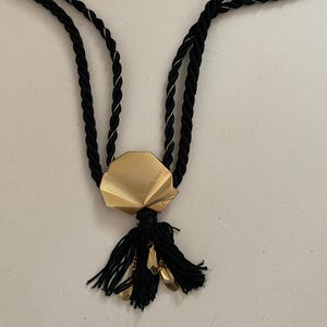 May include: A black rope necklace with a gold-colored geometric pendant and a black tassel. The pendant has a folded design and small gold-colored charms. The necklace is on a white background.