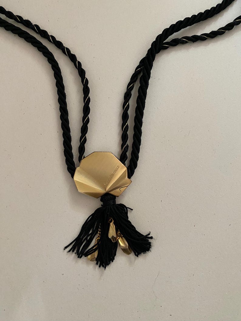 May include: A black rope necklace with a gold-colored geometric pendant and a black tassel. The pendant has a folded design and small gold-colored charms. The necklace is on a white background.