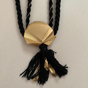 May include: A gold-toned pendant necklace featuring a shell-shaped charm with the engraved name "Gianni Versace." The necklace has a black rope-like cord with gold accents and a black tassel with gold-colored charms.