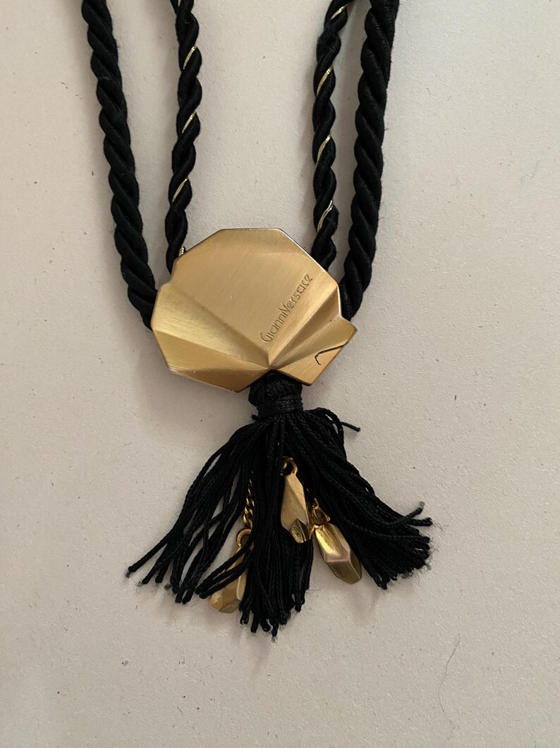 May include: A gold-toned pendant necklace featuring a shell-shaped charm with the engraved name "Gianni Versace." The necklace has a black rope-like cord with gold accents and a black tassel with gold-colored charms.