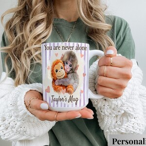 May include: White ceramic mug featuring a cartoon illustration of two monkeys hugging, with the text "You are never alone" and "Taylor's Mug" printed on the front. The mug has a white handle and is decorated with purple stripes and pink hearts.