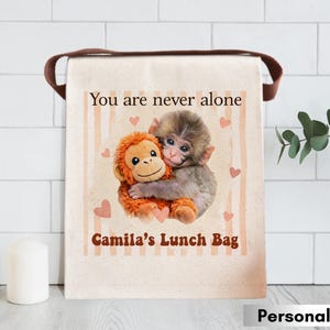 May include: A beige lunch bag with a brown handle features a graphic of two hugging monkeys, surrounded by hearts. The text "You are never alone" and "Camila's Lunch Bag" are printed on the bag. The bag is personalized.