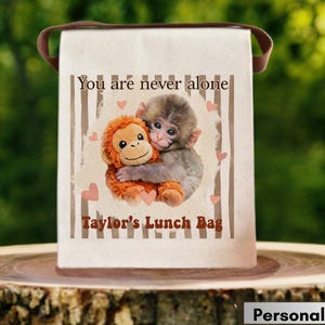 May include: A personalized lunch bag with a brown handle. The bag features a graphic of two monkeys hugging, with the text "You are never alone" and "Taylor's Lunch Bag". The background has vertical stripes and heart shapes.