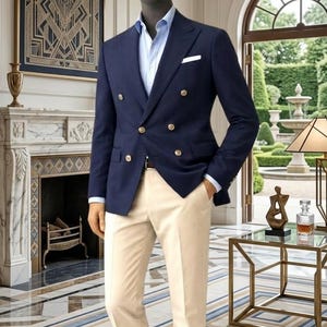 May include: A navy blue double-breasted blazer with gold buttons, paired with light beige pants and a light blue dress shirt. The mannequin is wearing brown leather shoes. The setting is a well-lit room with a decorative fireplace and a patterned rug.