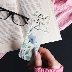 May include: A floral bookmark with the words "faith, hope, love" in elegant script. The bookmark features watercolor-style blue flowers and green leaves. The bookmark is held near an open book and glasses.