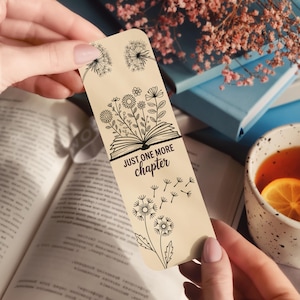Puede incluir: Un marcap&aacute;ginas beige con dibujos lineales negros de dientes de le&oacute;n, flores y un libro abierto. El texto "JUST ONE MORE chapter" est&aacute; impreso en el marcap&aacute;ginas. El marcap&aacute;ginas se sostiene sobre un libro abierto, con una taza de t&eacute; y libros en el fondo.