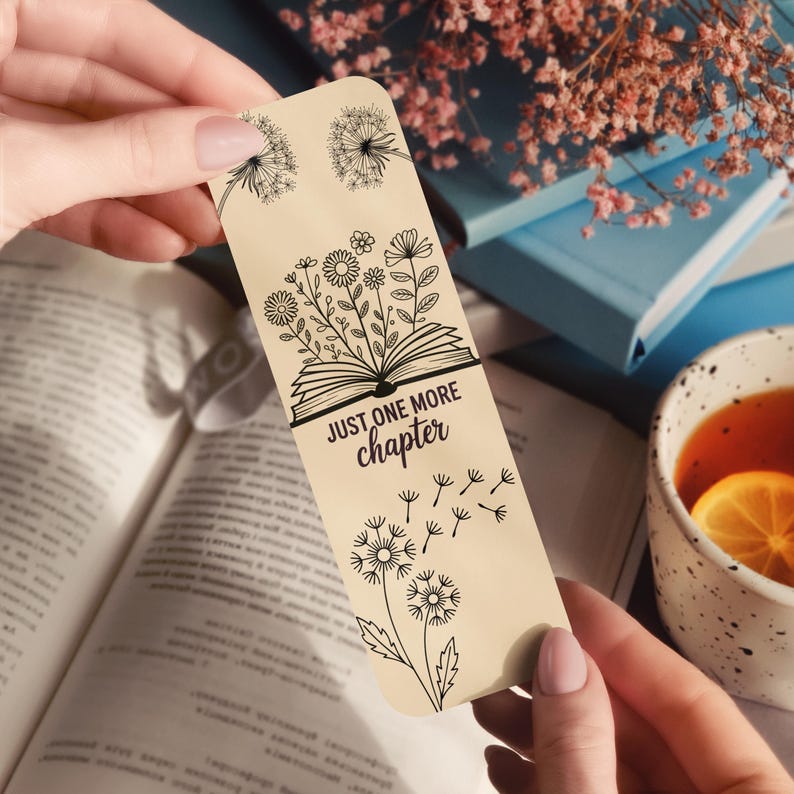 Puede incluir: Un marcap&aacute;ginas beige con dibujos lineales negros de dientes de le&oacute;n, flores y un libro abierto. El texto "JUST ONE MORE chapter" est&aacute; impreso en el marcap&aacute;ginas. El marcap&aacute;ginas se sostiene sobre un libro abierto, con una taza de t&eacute; y libros en el fondo.