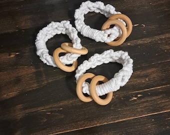 May include: Three white macrame baby teethers with natural wooden rings. The teethers are arranged on a dark brown wooden surface. Each teether has a circular design with a woven texture.
