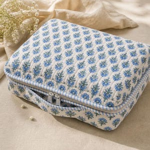 May include: A quilted, rectangular cosmetic bag with a blue floral pattern on a cream background. The bag has a zipper closure, a small handle, and a blue and white striped trim. The bag is on a cream-colored surface.