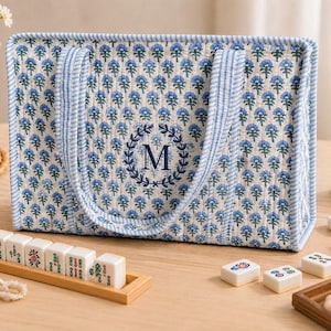 Blue floral quilted mahjong tote bag with striped handles, navy blue embroidered monogram “M” wreath detail, padded structure, and spacious interior for American mahjong tiles, displayed on a cozy tabletop with mahjong accessories.