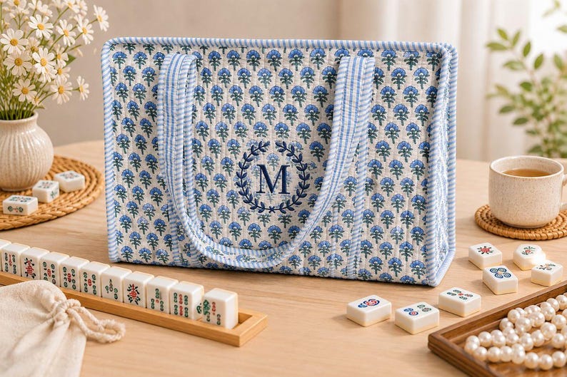 Blue floral quilted mahjong tote bag with striped handles, navy blue embroidered monogram “M” wreath detail, padded structure, and spacious interior for American mahjong tiles, displayed on a cozy tabletop with mahjong accessories.