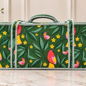 May include: A rectangular quilted storage bag featuring a dark green fabric with a repeating pattern of pink birds, yellow flowers, and pink butterflies. The bag has two top handles and white and green striped piping along all edges.