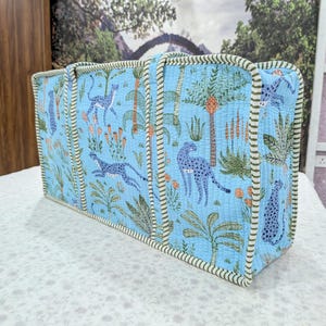 May include: A rectangular quilted tote bag in light blue featuring a repeating pattern of blue leopards, green palm trees, and orange floral elements. The bag has two vertical handles and is finished with a green and white striped fabric trim.
