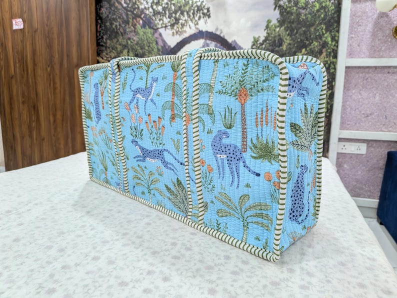 May include: A rectangular quilted tote bag in light blue featuring a repeating pattern of blue leopards, green palm trees, and orange floral elements. The bag has two vertical handles and is finished with a green and white striped fabric trim.