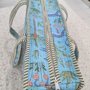 May include: A light blue quilted tote bag featuring a repeating botanical print of green leaves, orange flowers, and blue abstract shapes. The bag has a white zipper closure and is finished with green and white striped fabric trim and two handles.