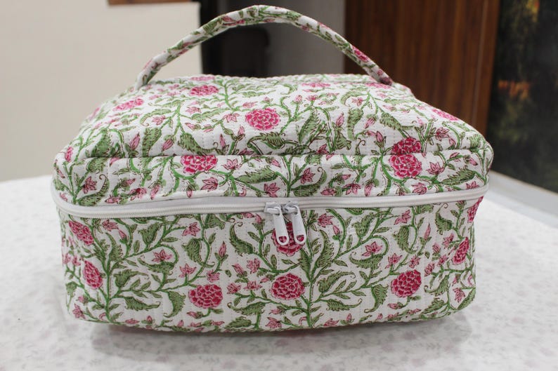 May include: A floral print cosmetic bag with a white zipper and handle. The bag features a pattern of pink flowers and green leaves on a white background. The bag is rectangular with rounded edges.