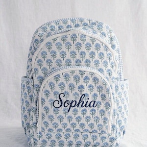 以下が含まれることがあります： 青い花柄のキルティングバックパックで、前面に「Sophia」という名前が刺繍されています。 バックパックには白いジッパーとサイドポケットが付いています。 背景は明るくニュートラルな色です。