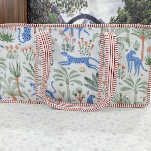 May include: A rectangular quilted tote bag featuring a white background with a repeating pattern of blue leopards, green palm trees, and orange flowers. The bag has two handles and edges trimmed with a red and white striped fabric pattern.