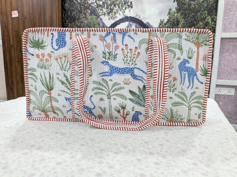 May include: A rectangular quilted tote bag featuring a white background with a repeating pattern of blue leopards, green palm trees, and orange flowers. The bag has two handles and edges trimmed with a red and white striped fabric pattern.