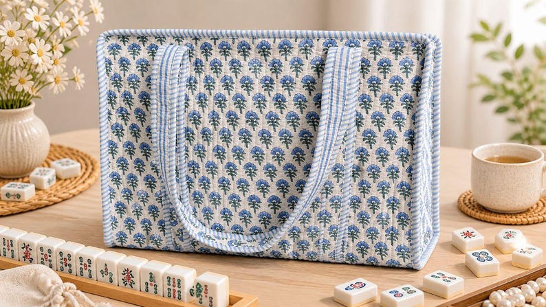 May include: A rectangular quilted tote bag featuring a repeating blue and green floral block print pattern on a white background, finished with light blue and white striped fabric trim along the edges and two matching striped shoulder straps.