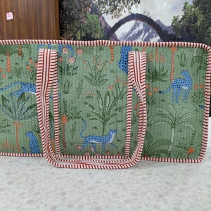 May include: A rectangular quilted tote bag in sage green featuring a repeating pattern of blue cheetahs and tropical plants. The bag is finished with red and white striped fabric trim along the edges and includes two matching striped shoulder straps.