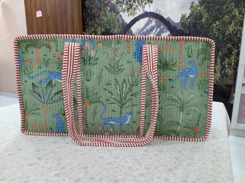 May include: A rectangular quilted tote bag in sage green featuring a repeating pattern of blue cheetahs and tropical plants. The bag is finished with red and white striped fabric trim along the edges and includes two matching striped shoulder straps.