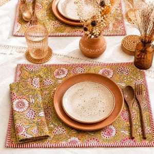 Può includere: Un'apparecchiatura da tavola con tovagliette, tovaglioli e piatti a motivi floreali. Le tovagliette sono gialle con disegni floreali rosa e bianchi e bordi rossi. Sono presenti anche posate di legno e un bicchiere d'acqua.