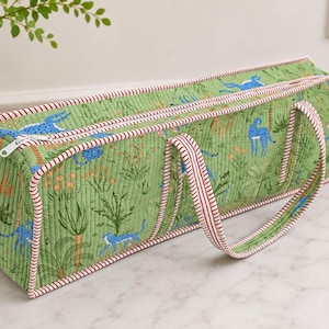 May include: A rectangular quilted fabric bag featuring a green background with a repeating pattern of blue cheetahs and tropical plants. The bag has a white zipper closure and red and white striped piping along the edges and handles.