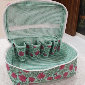 May include: Open, floral-patterned cosmetic bag with a white zipper and interior pockets. The bag features a green and white striped interior and a floral design with pink flowers and green leaves on a teal background.