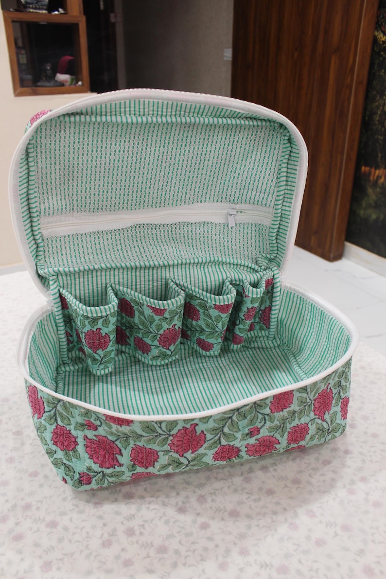 May include: Open, floral-patterned cosmetic bag with a white zipper and interior pockets. The bag features a green and white striped interior and a floral design with pink flowers and green leaves on a teal background.