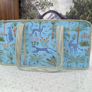 May include: A rectangular quilted tote bag in light blue featuring a repeating pattern of dark blue cheetahs, palm trees, and orange flowers. The bag has two long, striped fabric handles and matching striped piping along all the outer edges.