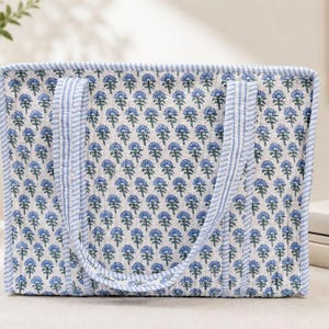 May include: A rectangular quilted tote bag featuring a repeating blue floral block print pattern on a white background. The bag has two matching fabric handles and is finished with a blue and white striped trim along the edges and seams.