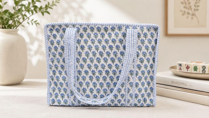 May include: A rectangular quilted tote bag featuring a repeating blue floral block print pattern on a white background. The bag has two matching fabric handles and is finished with a blue and white striped trim along the edges and seams.