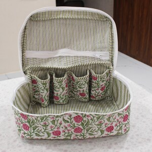 May include: Open, white cosmetic bag with a floral print. The bag has a white zipper and a striped interior. The interior has four small pockets with a floral design. The exterior has a floral pattern with pink flowers and green leaves.