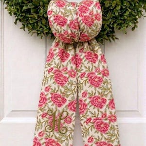 K&ouml;nnte beinhalten: Eine dekorative Stoffschleife mit einem Blumenmuster aus rosa Rosen und gr&uuml;nen Bl&auml;ttern auf cremefarbenem Grund. Die Schleife hat zwei lange, herabh&auml;ngende B&auml;nder, wobei das linke Band mit einem gro&szlig;en, kursiven Buchstaben H in Olivgr&uuml;n bestickt ist.