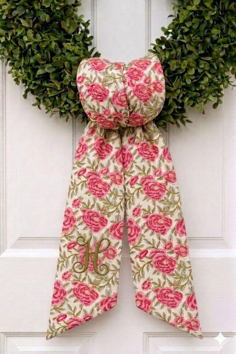 K&ouml;nnte beinhalten: Eine dekorative Stoffschleife mit einem Blumenmuster aus rosa Rosen und gr&uuml;nen Bl&auml;ttern auf cremefarbenem Grund. Die Schleife hat zwei lange, herabh&auml;ngende B&auml;nder, wobei das linke Band mit einem gro&szlig;en, kursiven Buchstaben H in Olivgr&uuml;n bestickt ist.